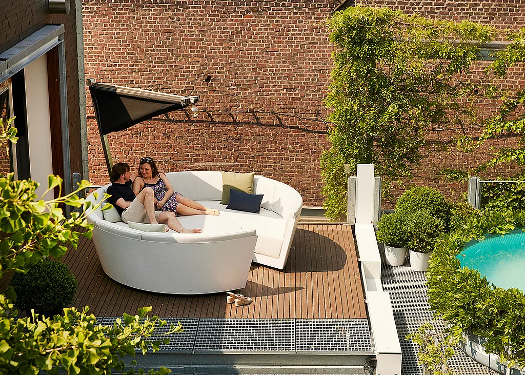 White Kosmos se prélasse sur une terrasse en bois sur le toit, où un couple s’assoit ensemble sous une ombre pliante noire à côté d’un mur de briques, escaladant de la verdure et un aperçu d’une piscine bleue.