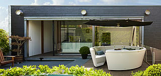 Weite Aussicht auf die Dachterrasse mit weißer Kosmos-Lounge und schwarzem Schatten vor einer dunklen modernen Fassade mit großen Glasschiebetüren