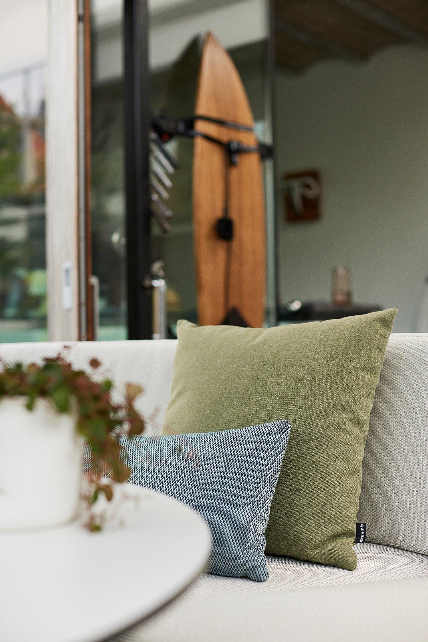 Nahaufnahme der grünen und blauen Kissen auf dem weißen Kosmos-Sofa, mit einer offenen Glastür und einem im Haus dahinter sichtbaren hölzernen Surfbrett.