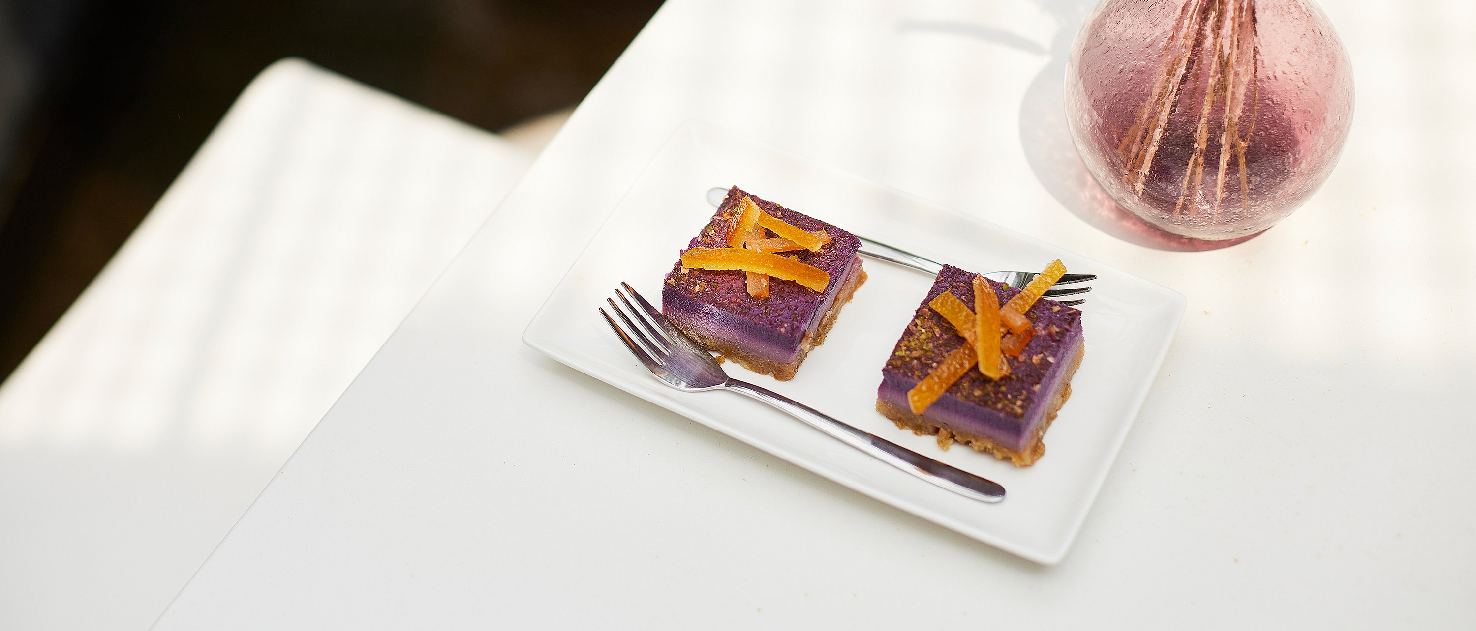 Nahaufnahme von zwei violetten Gebäckstücken mit Orangenschale auf einem weißen Teller neben einer rosa Glasvase auf der weißen Picnik-Tischplatte.
