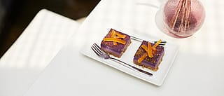 Nahaufnahme von zwei violetten Gebäckstücken mit Orangenschale auf einem weißen Teller neben einer rosa Glasvase auf der weißen Picnik-Tischplatte.