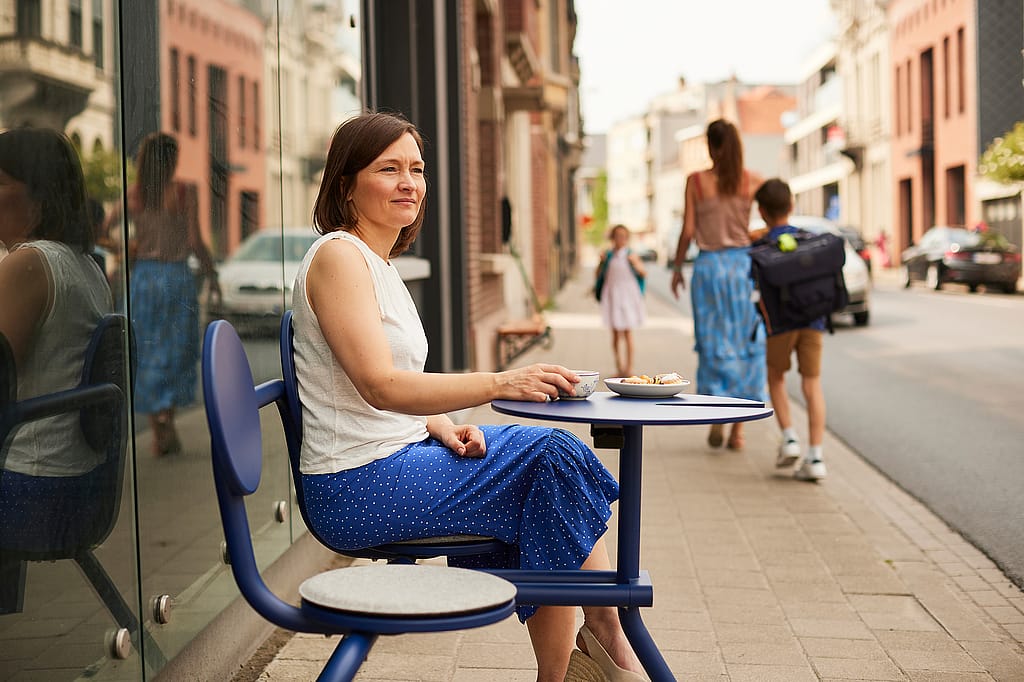 Frau sitzt auf einem kobaltblauen Bistroo-Stuhl mit Tisch auf einem Bürgersteig in der Stadt, mit einer Tasse und einem Teller auf der Tischplatte und Fußgängern, die hinter ihr die Straße entlanggehen.