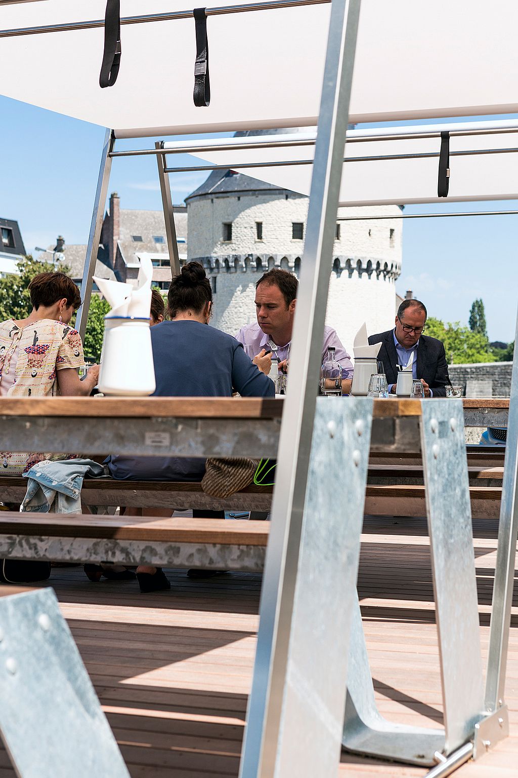 People dining together at a Hopper picnic table under a white canopy, framed by galvanized table legs, with the historic tower visible behind them.
