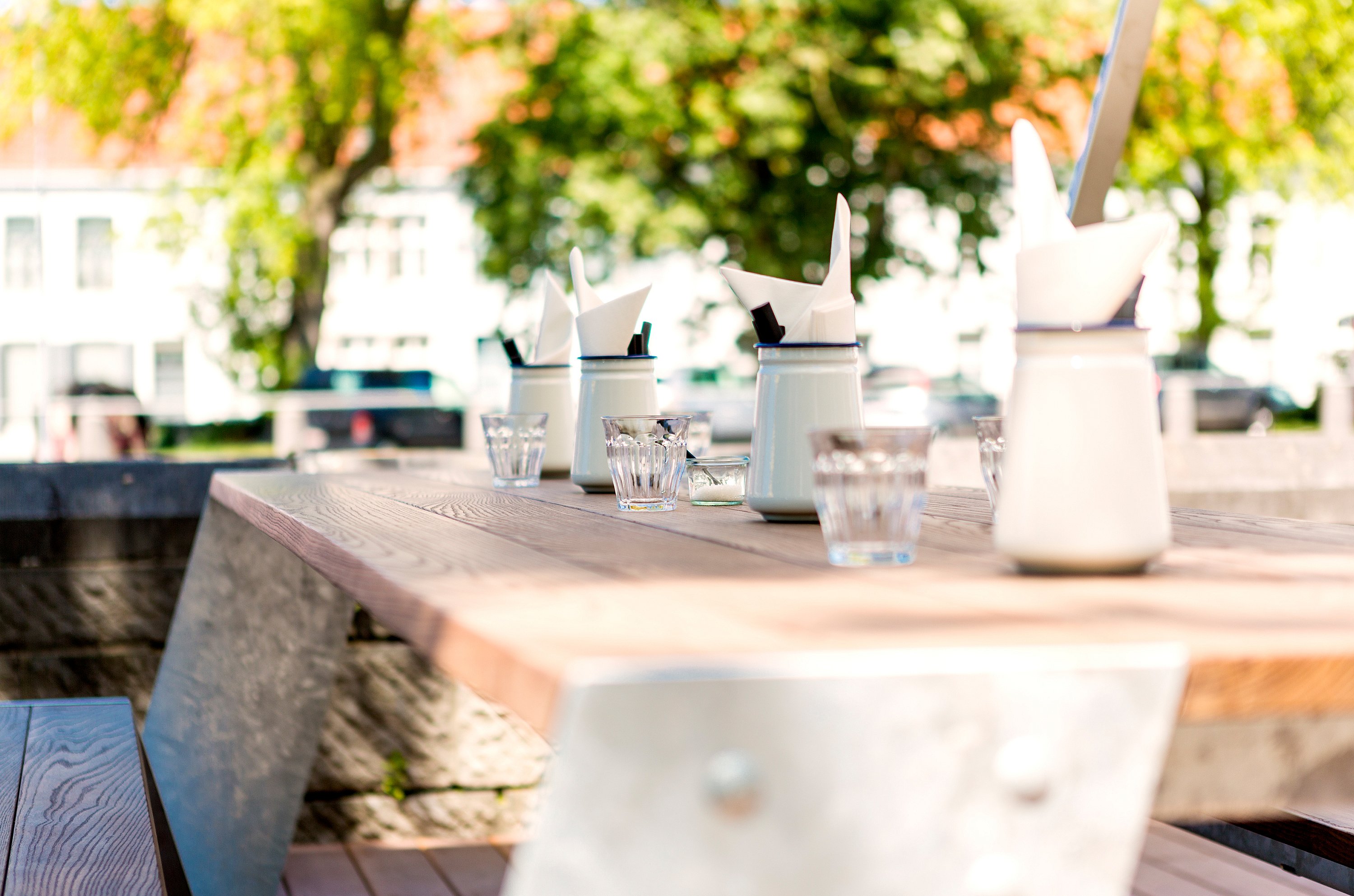 Close-up of a wooden Hopper tabletop set with white condiment pots, folded napkins and small drinking glasses, with sunlit trees softly blurred in the background.