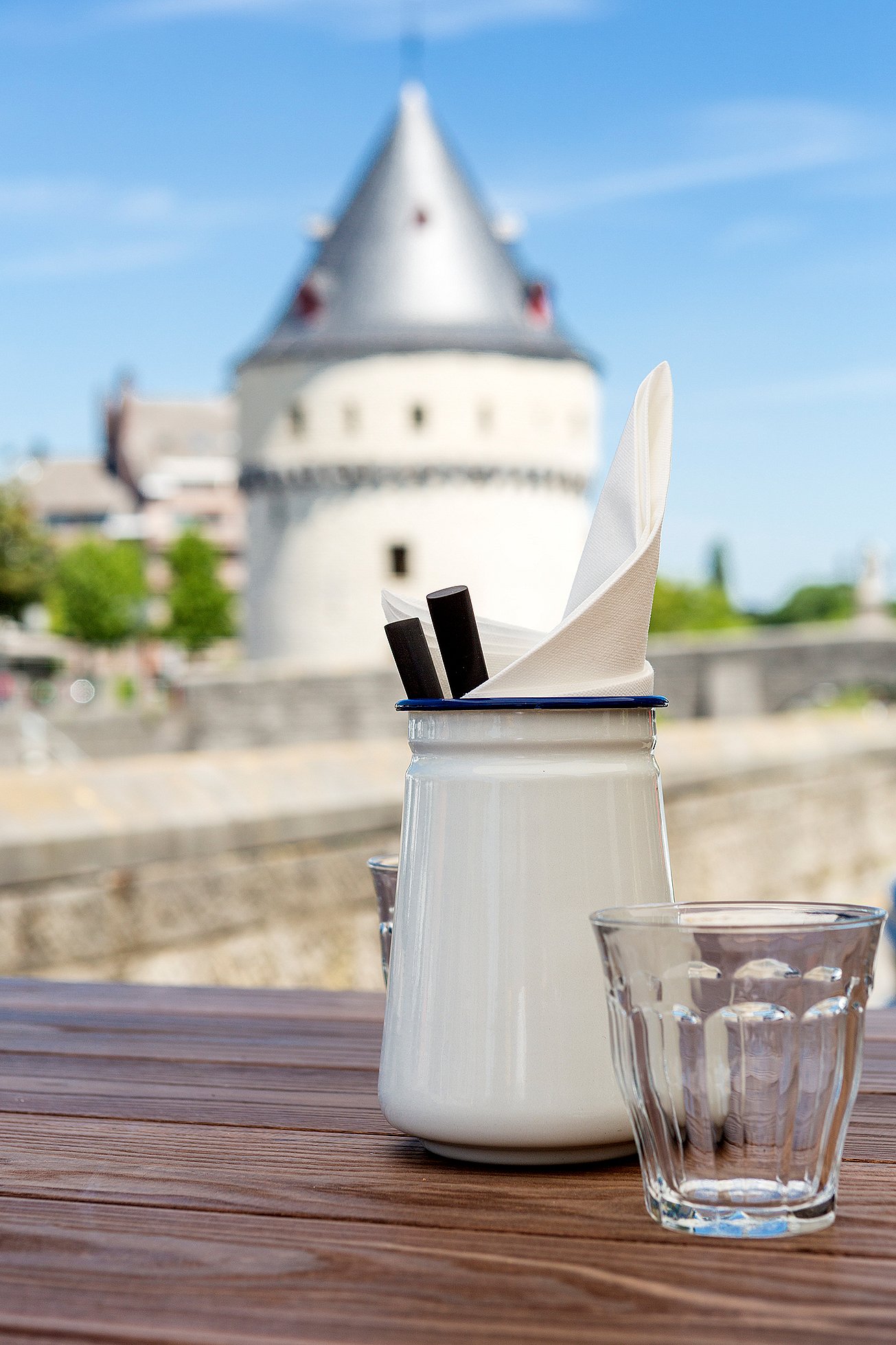 White condiment pot and glass on a wooden outdoor table, with a round stone tower softly blurred across the river in the background.