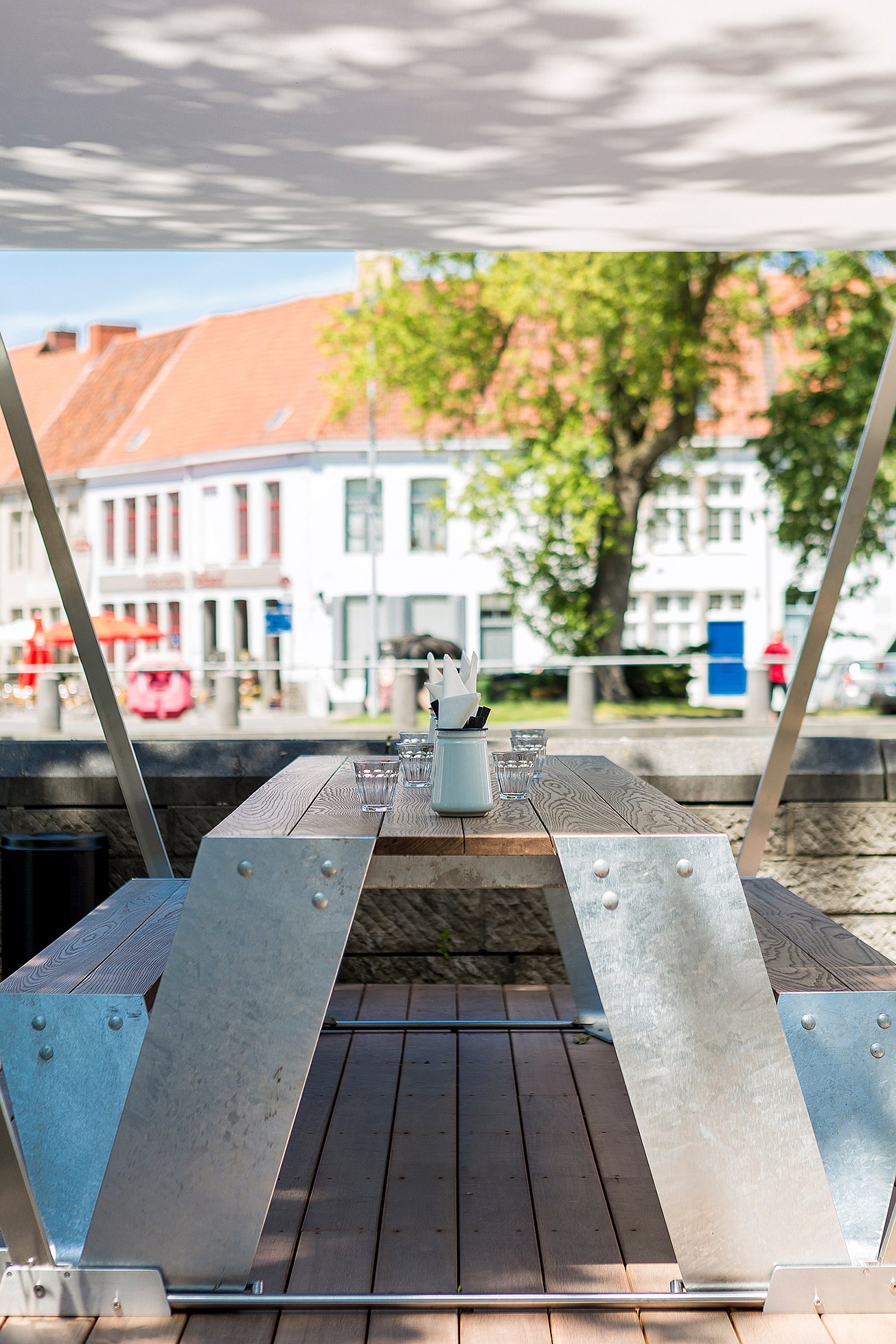 Empty Hopper picnic table centered under a white canopy on a wooden terrace, set with glasses and condiments and facing the riverside street beyond.
