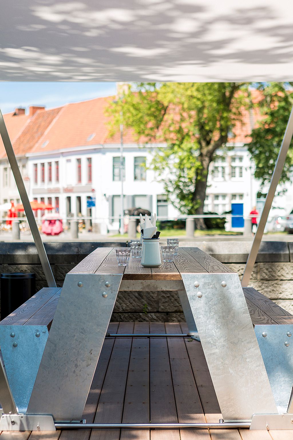 Empty Hopper picnic table centered under a white canopy on a wooden terrace, set with glasses and condiments and facing the riverside street beyond.