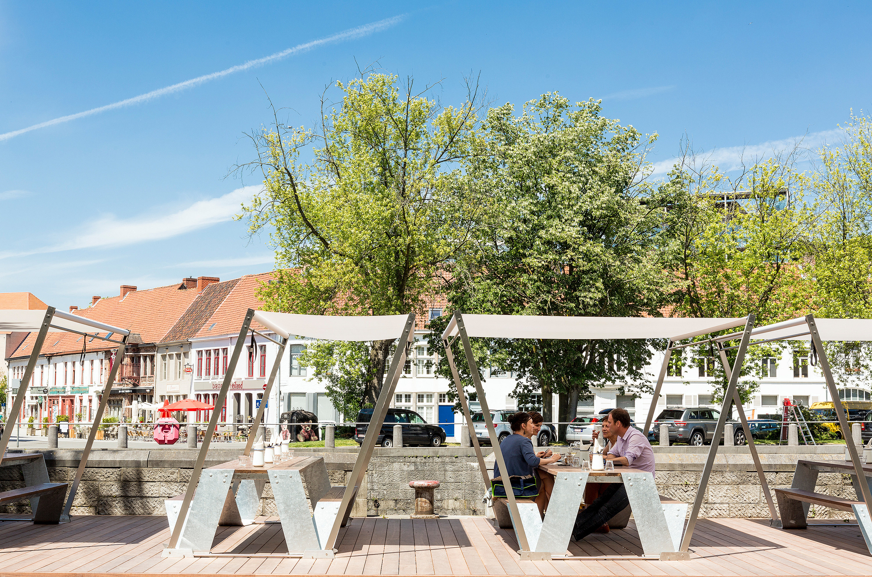 Picknicktafels van het merk Hopper met witte schaduwdoeken op een houten terras aan de rivier, waar gasten aan de oever van de Leie zitten met bomen en historische gebouwen aan de overkant.