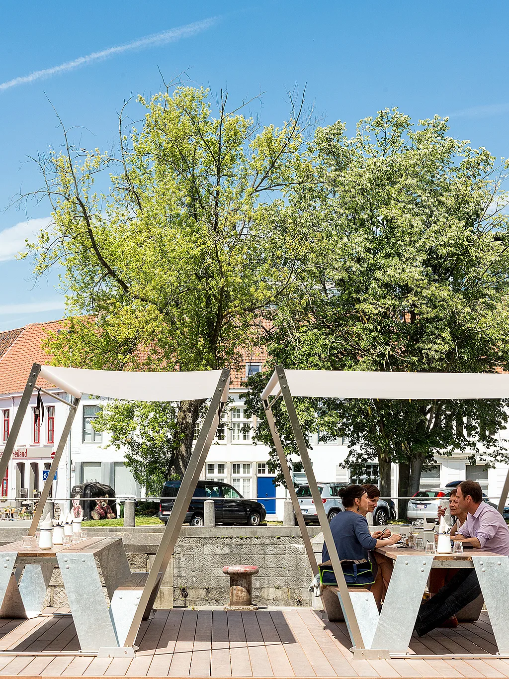 Picknicktafels van het merk Hopper met witte schaduwdoeken op een houten terras aan de rivier, waar gasten aan de oever van de Leie zitten met bomen en historische gebouwen aan de overkant.