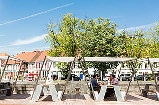 Hopper picnic tables with white shade canopies on a wooden riverside terrace, where diners sit beside the Leie with trees and historic buildings across the water.