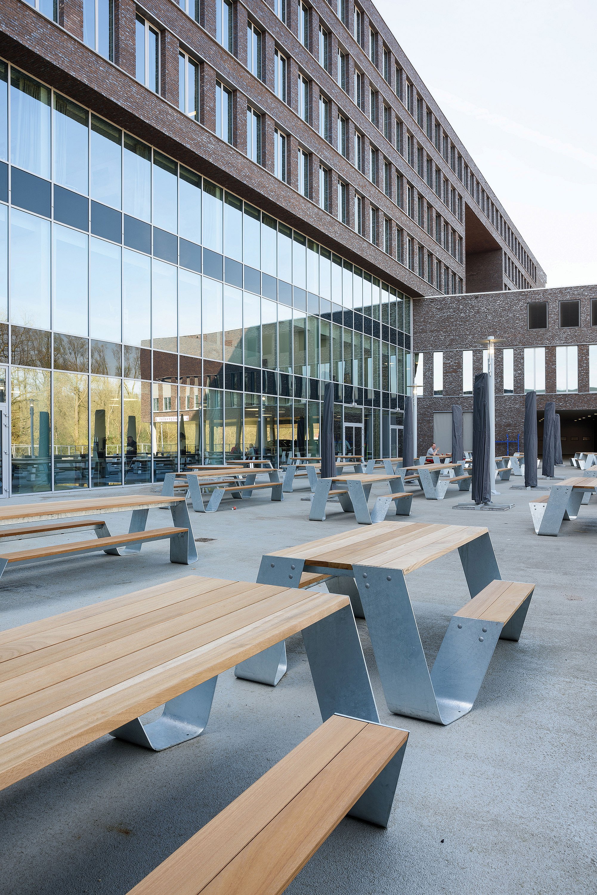 Plusieurs tables de pique-nique Hopper avec des pieds galvanisés et des dessus en bois répartis sur une spacieuse terrasse d’hôpital à côté d’une grande façade en verre et d’un bâtiment moderne en brique.