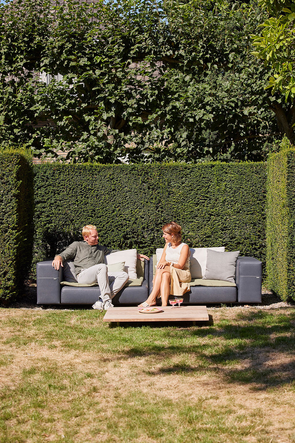 Mann und Frau unterhalten sich auf einem dunkelgrauen Walrus-Lounge-Sofa in einem gepflegten Garten, mit einem niedrigen Holztisch und ordentlich abgeschnittenen Hecken, die die Szene einrahmen.