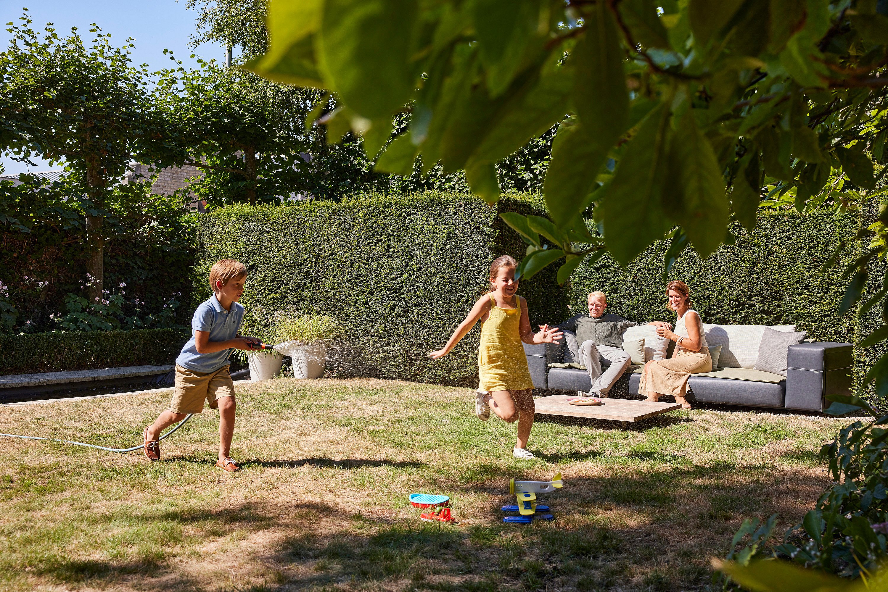 Zwei Kinder rennen über einen sonnigen Gartenrasen, eines hält einen Schlauch, während Erwachsene im Hintergrund auf dem Walrus-Sofa entspannen.