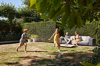 Zwei Kinder rennen über einen sonnigen Gartenrasen, eines hält einen Schlauch, während Erwachsene im Hintergrund auf dem Walrus-Sofa entspannen.