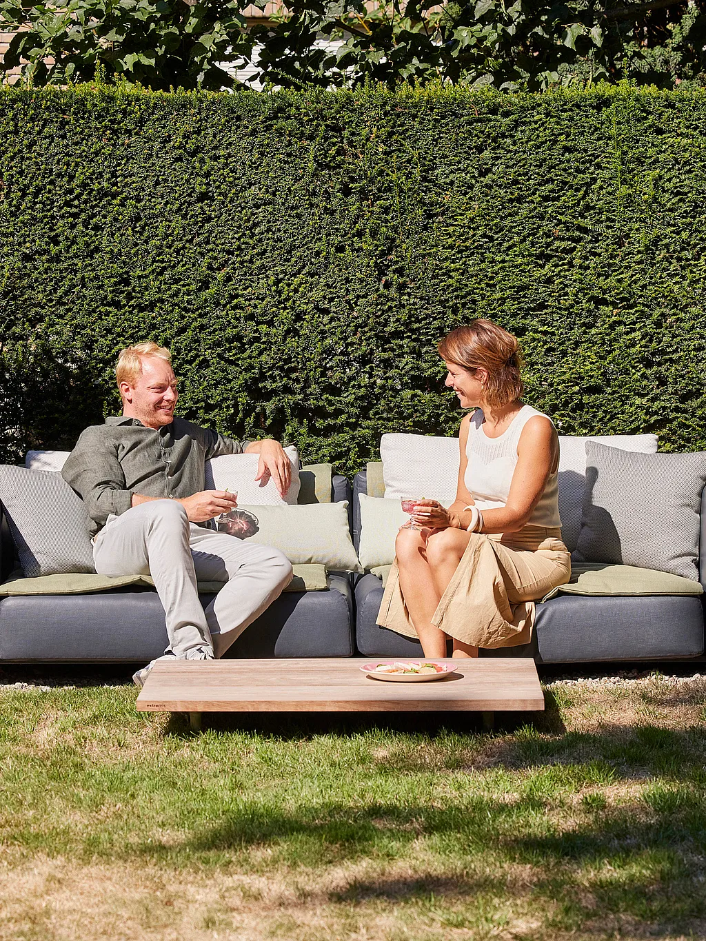 Koppel zittend op een donkergrijze Walrus buitenbank in een netjes bijgesneden tuin, met een lage houten salontafel vooraan en hoge geknipte heggen erachter.