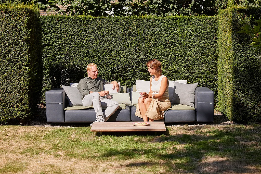 Paar sitzt auf einem dunkelgrauen Walross-Außensofa in einem ordentlich gepflegten Garten, mit einem niedrigen Couchtisch aus Holz vorne und hohen abgeschnittenen Hecken dahinter.