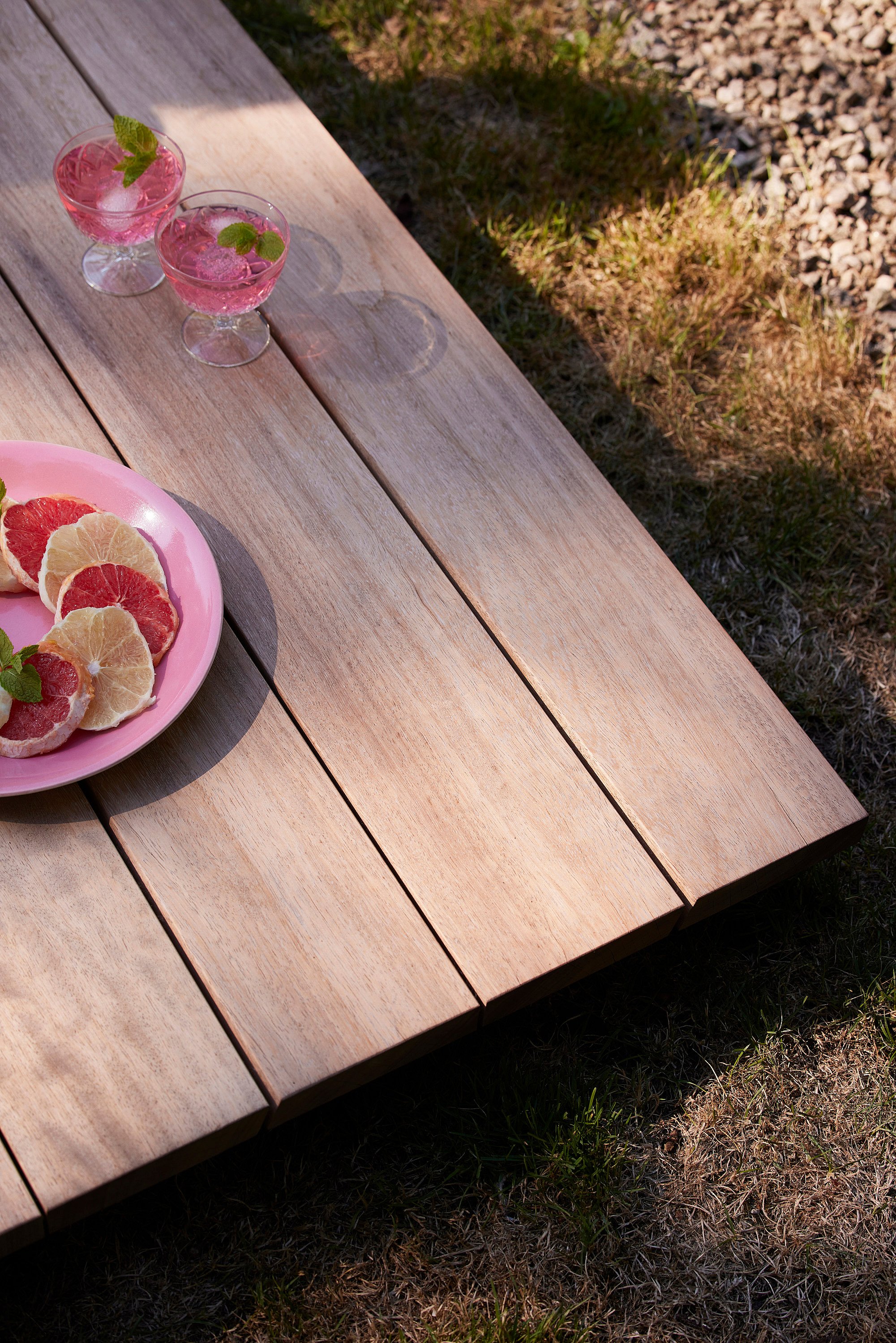 Niedriger Couchtisch aus Holz im Freien mit einem rosa Teller geschnittener Grapefruit und zwei rosa Getränken, auf trockenem Gras neben der Lounge aufgestellt.