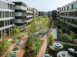 Blick von oben auf den Gemeinschaftshof mit Backsteinwegen, bepflanzten Beeten, Bänken, langen Gemeinschaftstischen und zwei runden Pantagruel-Picknicktischen inmitten der Bepflanzung.