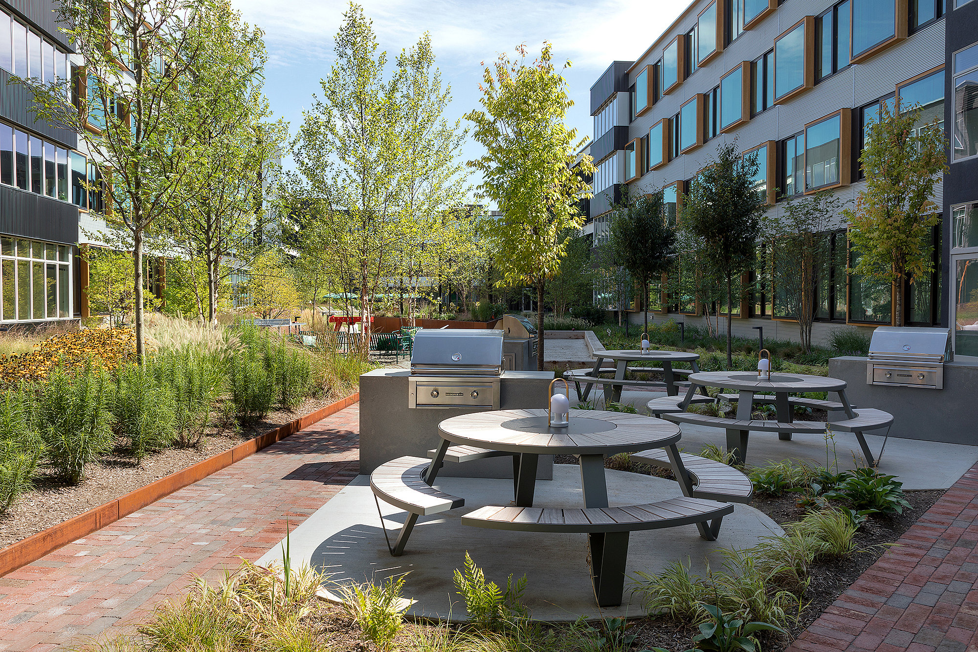 Landscaped courtyard between modern apartment buildings, with several gray Pantagruel picnic tables, built-in barbecue grills, brick walkways, and young trees in bright sunlight.