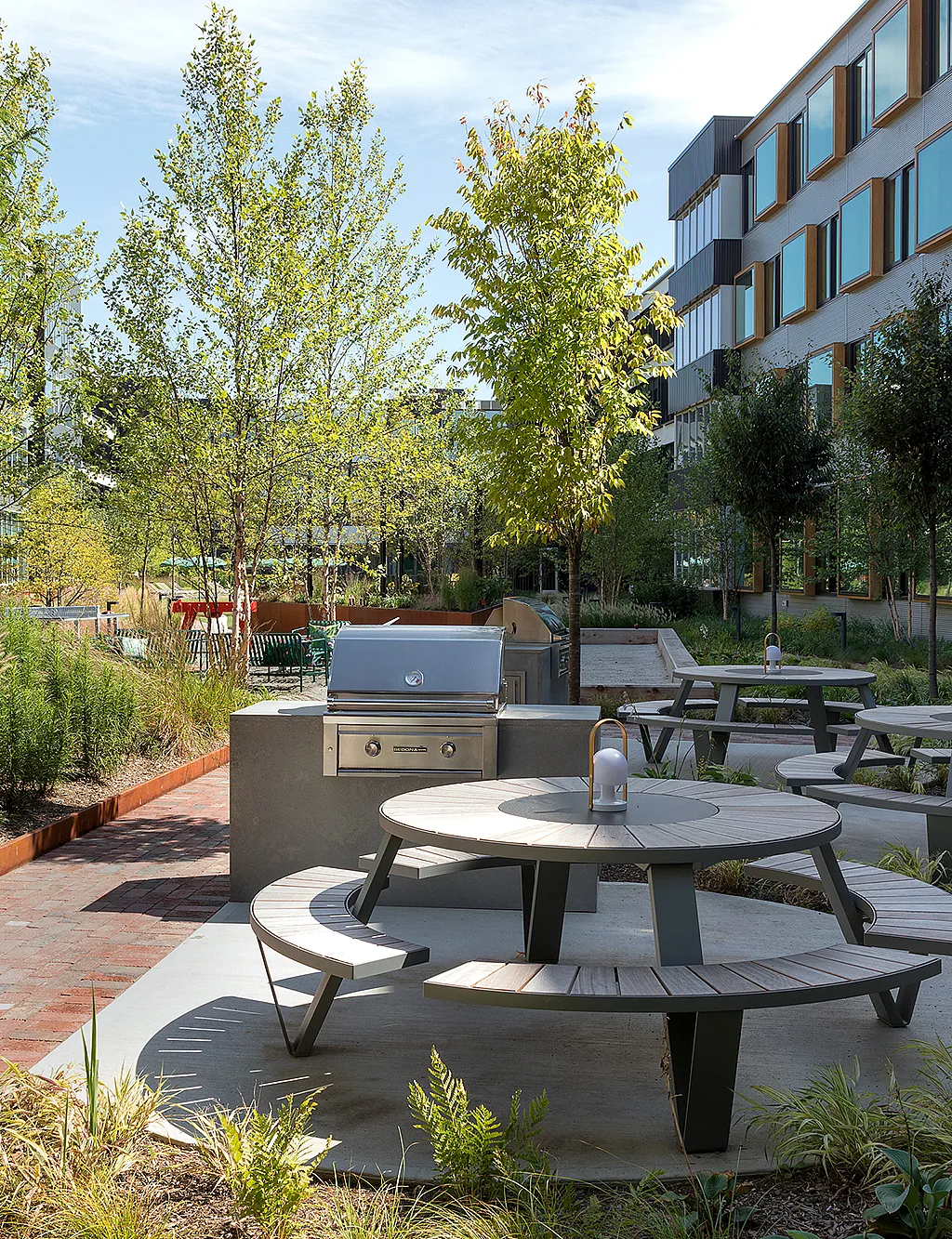 Landscaped courtyard between modern apartment buildings, with several gray Pantagruel picnic tables, built-in barbecue grills, brick walkways, and young trees in bright sunlight.