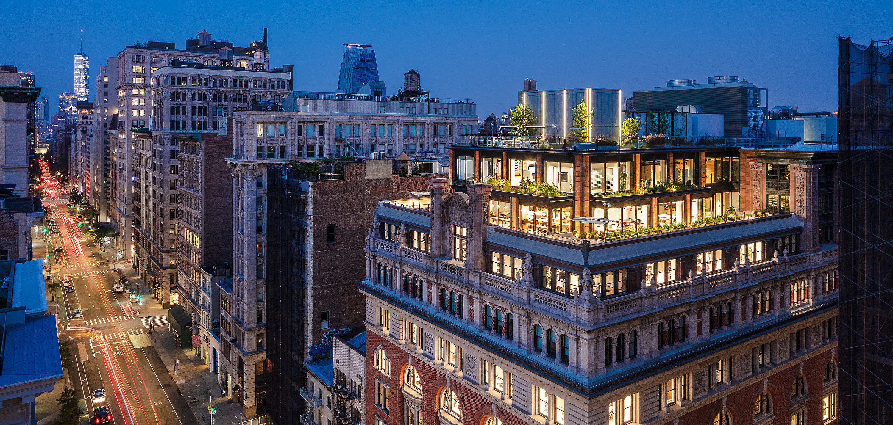 Abendlicher Stadtblick auf das beleuchtete Dachgebäude des Mastercard Tech Hub mit mehreren bepflanzten Terrassen und Sitzgelegenheiten im Freien über einer belebten Straße in Manhattan.