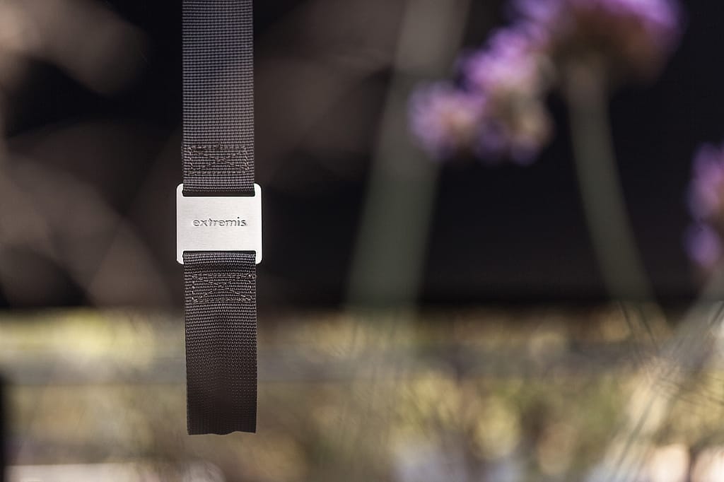 Close-up of a black hanging strap with a metal Extremis buckle beneath a shade canopy, with soft-focus flowers and table legs in the background.