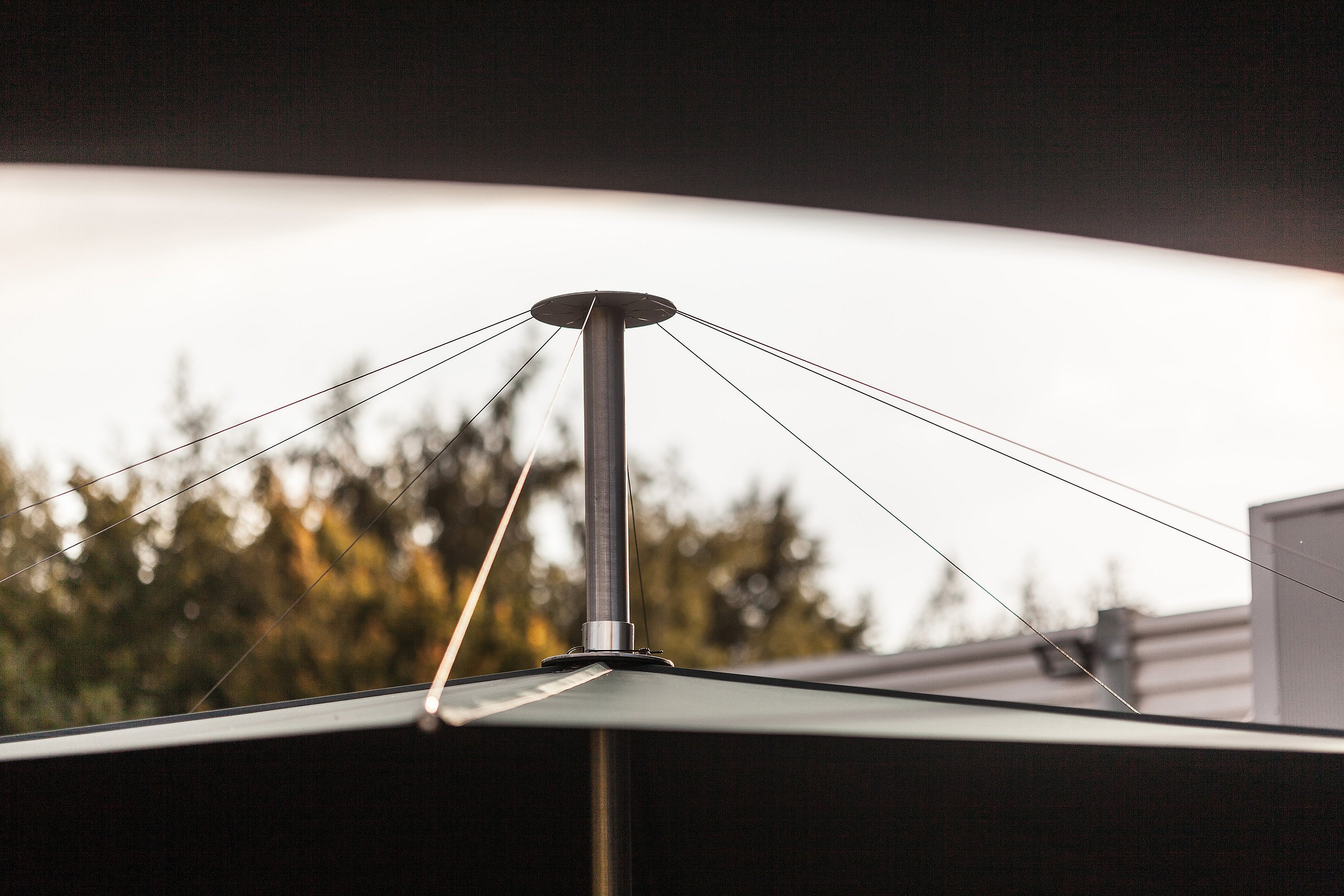 Close-up of the top of a black parasol, showing the central pole and tension cables against a pale sky and blurred trees.