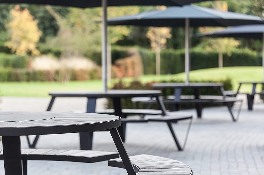 Close-up of a black round picnic table with curved benches in the foreground, with more black parasols and picnic tables softly blurred across the paved terrace behind.
