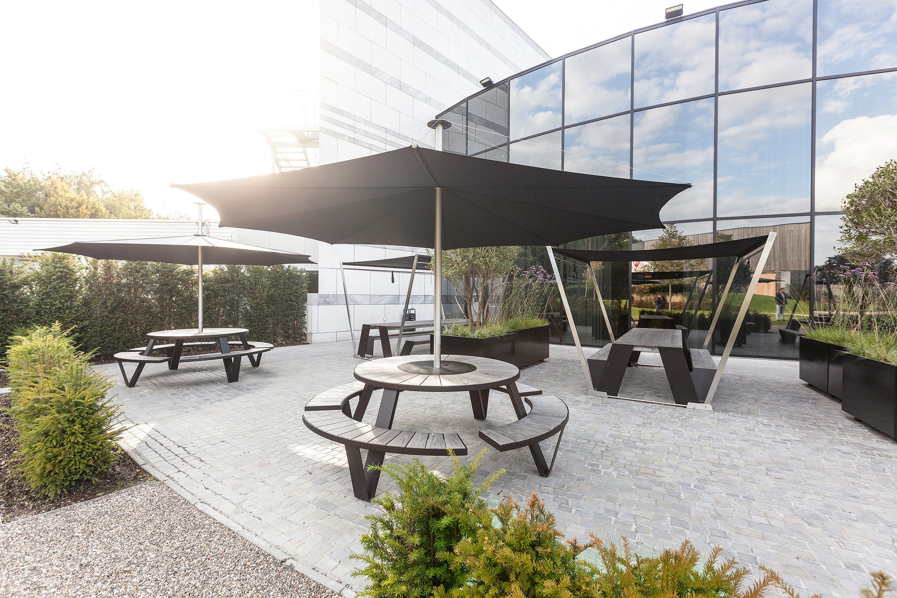 Landscaped courtyard outside a modern glass office building, with a pantagruel under a large black parasol in the foreground and a long black picnic table with matching shade beside reflective windows and planters.