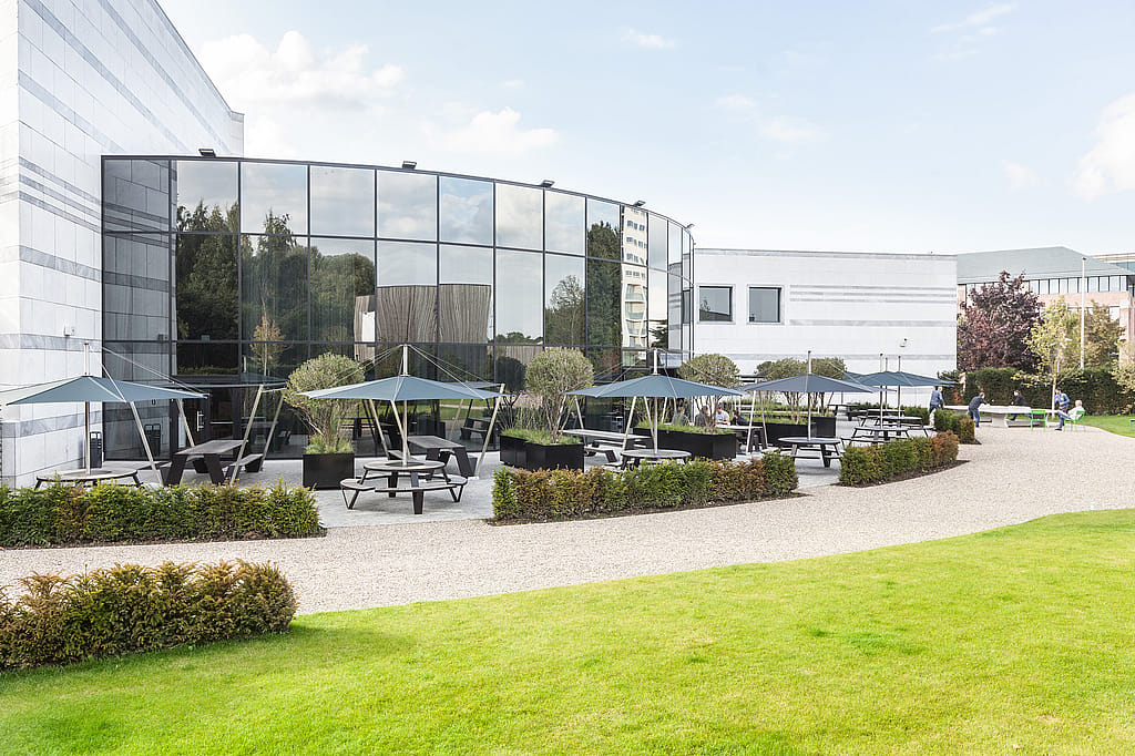 Broad exterior view of the broadcasting company’s grounds, with mirrored office façades, lawns, gravel walkways, and several shaded black pantagruel tables spread across the terrace.