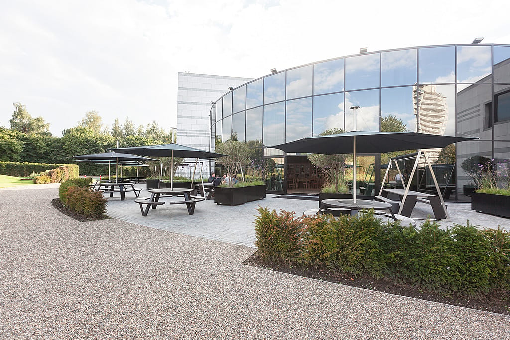 Wide view of the office terrace with several black pantagruels and matching inumbra parasols arranged along a curved mirrored façade, bordered by gravel paths, hedges, and planted containers.