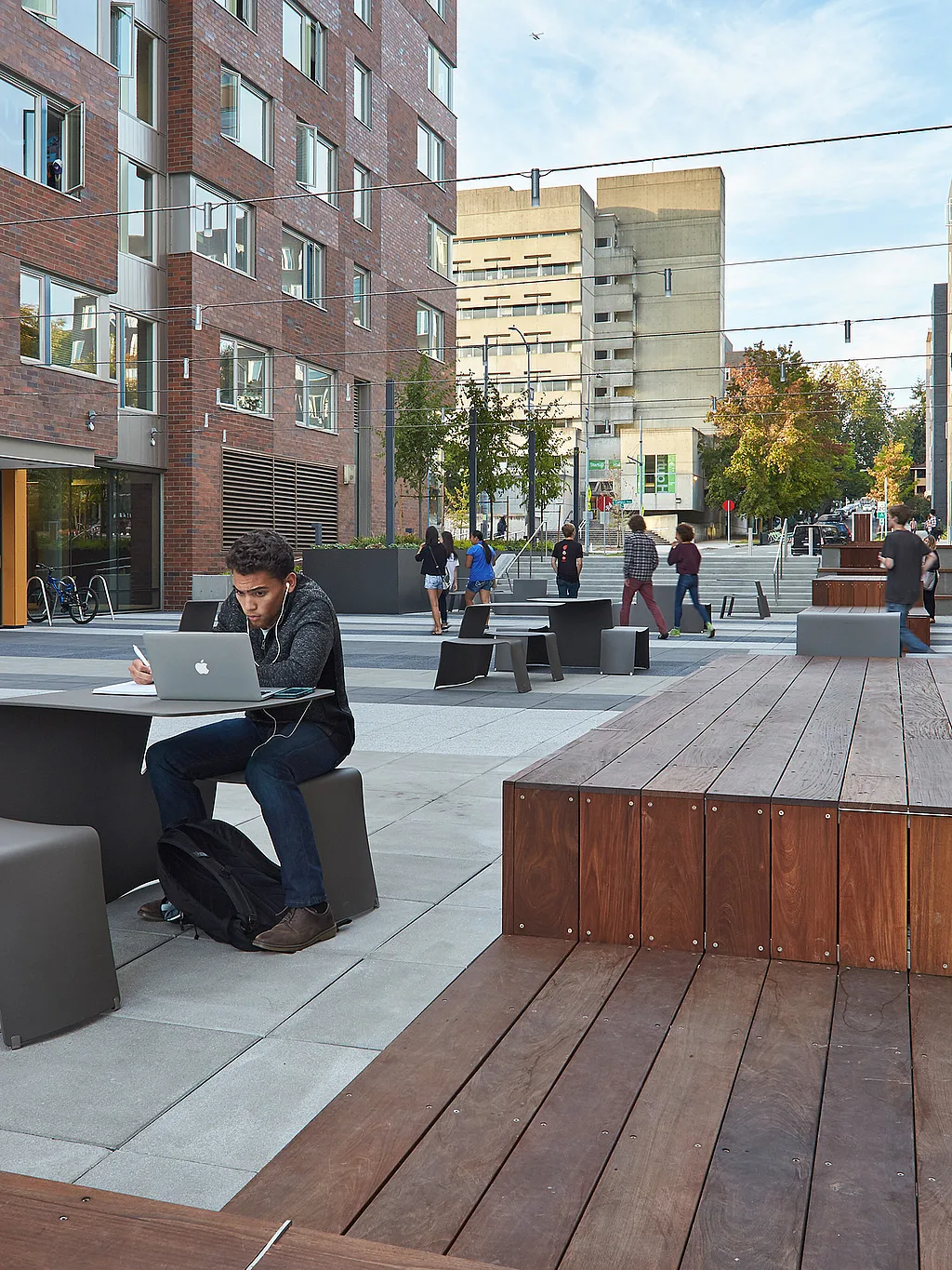 Student seated at a gray outdoor table in a broad paved plaza between modern residence halls, with wooden benches, overhead string lights, and groups of people walking and gathering in the background.