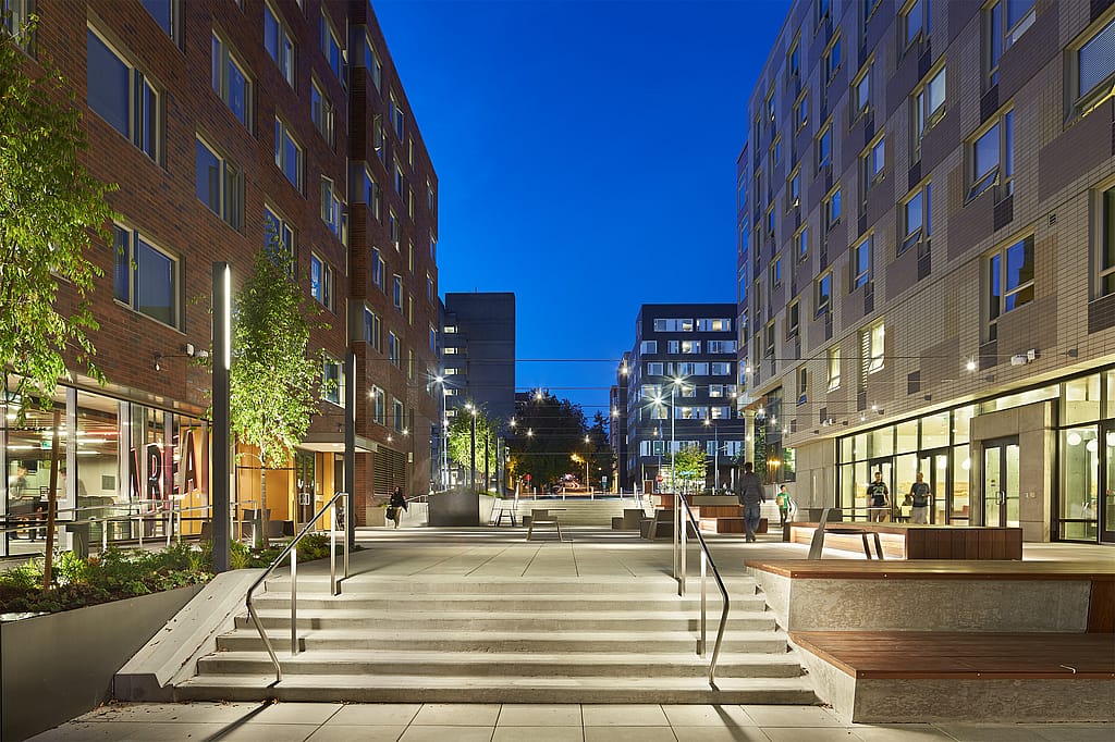 Vue en soirée de la place étudiante entre les résidences universitaires illuminées. On y voit de larges marches, de longs bancs en bois, des sièges extérieurs gris et quelques personnes qui se déplacent dans la cour éclairée.