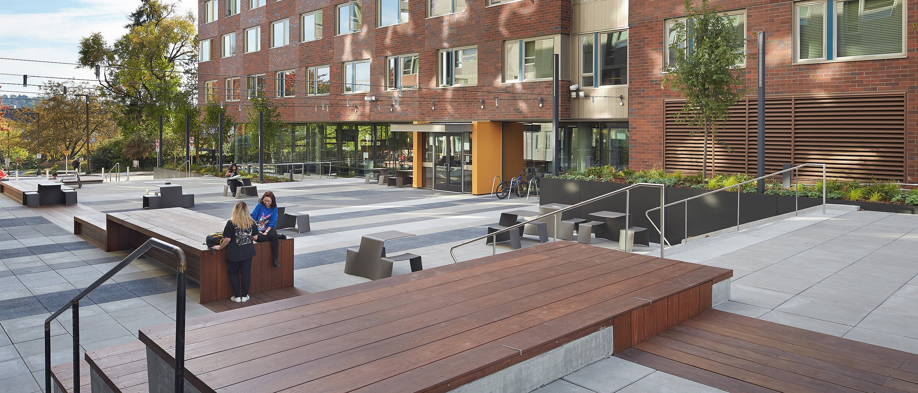   Vue d'ensemble d'une cour étudiante paysagée avec des terrasses en gradins, de longues estrades en bois, des tables et des tabourets gris en terrasse. Deux personnes discutent devant une résidence universitaire en briques.