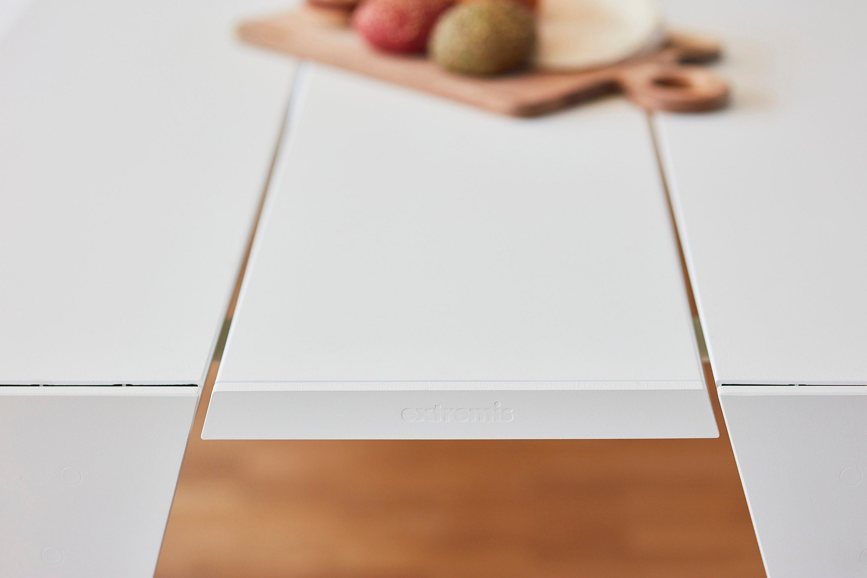 Close-up van de centrale witte tafelplank met het Extremis-logo, omlijst door de omringende tafeloppervlakken en een wazig serveerbord op de achtergrond.