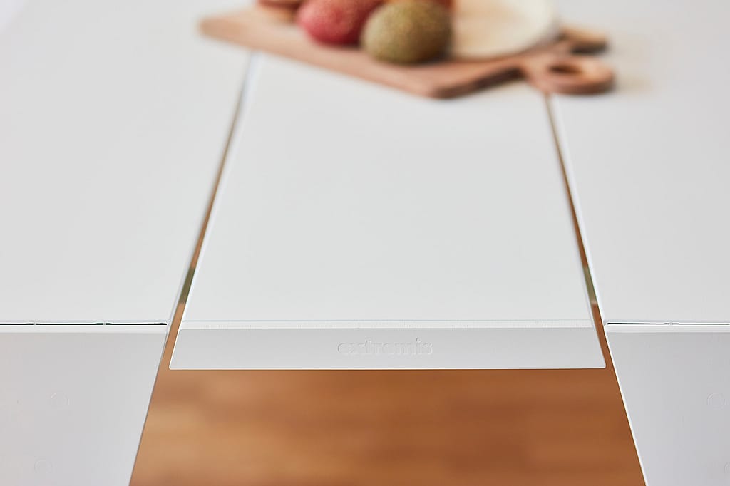 Close-up van de centrale witte tafelplank met het Extremis-logo, omlijst door de omringende tafeloppervlakken en een wazig serveerbord op de achtergrond.