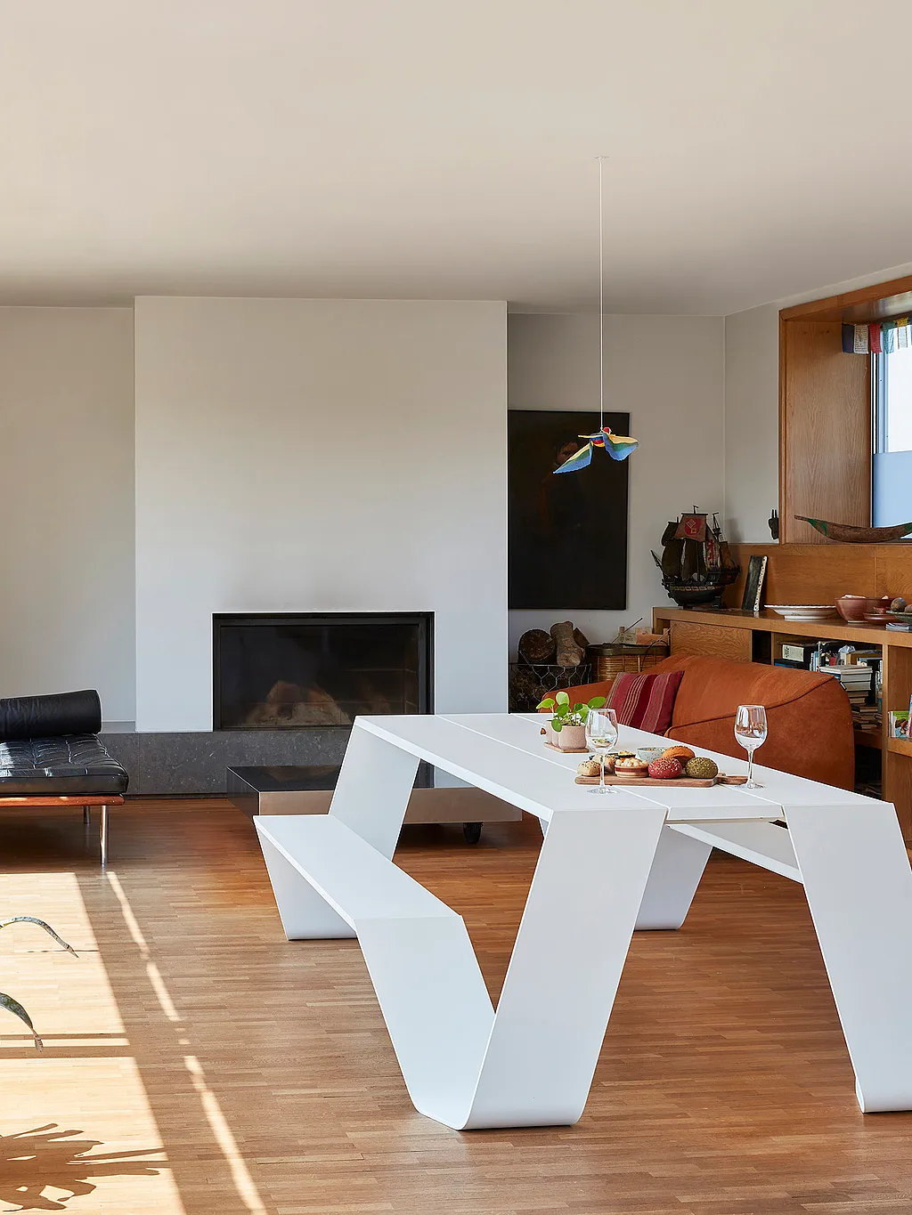 Salon minimaliste avec une table intérieure Hopper blanche et un banc sur un plancher en bois, à côté de fenêtres pleine hauteur, étagères encastrées et une cheminée noire.