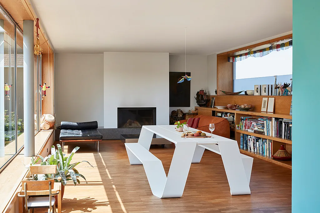 Minimalist living room with a white Hopper indoor table and bench on a wooden floor, beside full-height windows, built-in bookshelves, and a black fireplace.