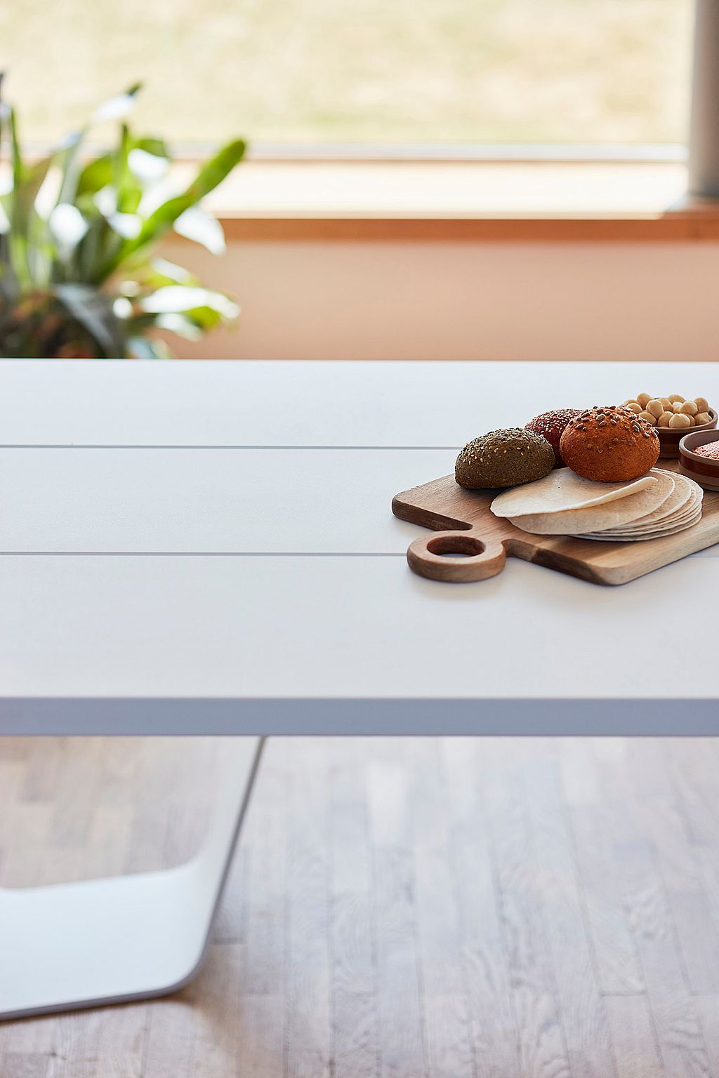 Wit tafelblad met een houten serveerbord met brood en kleine kommen, gefotografeerd bij zacht daglicht bij een raam.