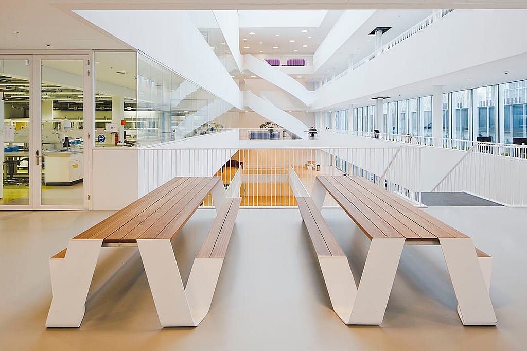 Vooraanzicht van twee lange Hopper picknicktafels met houten latten en witte poten, geplaatst naast een laboratoriumingang en met uitzicht op de hal met meerdere verdiepingen.