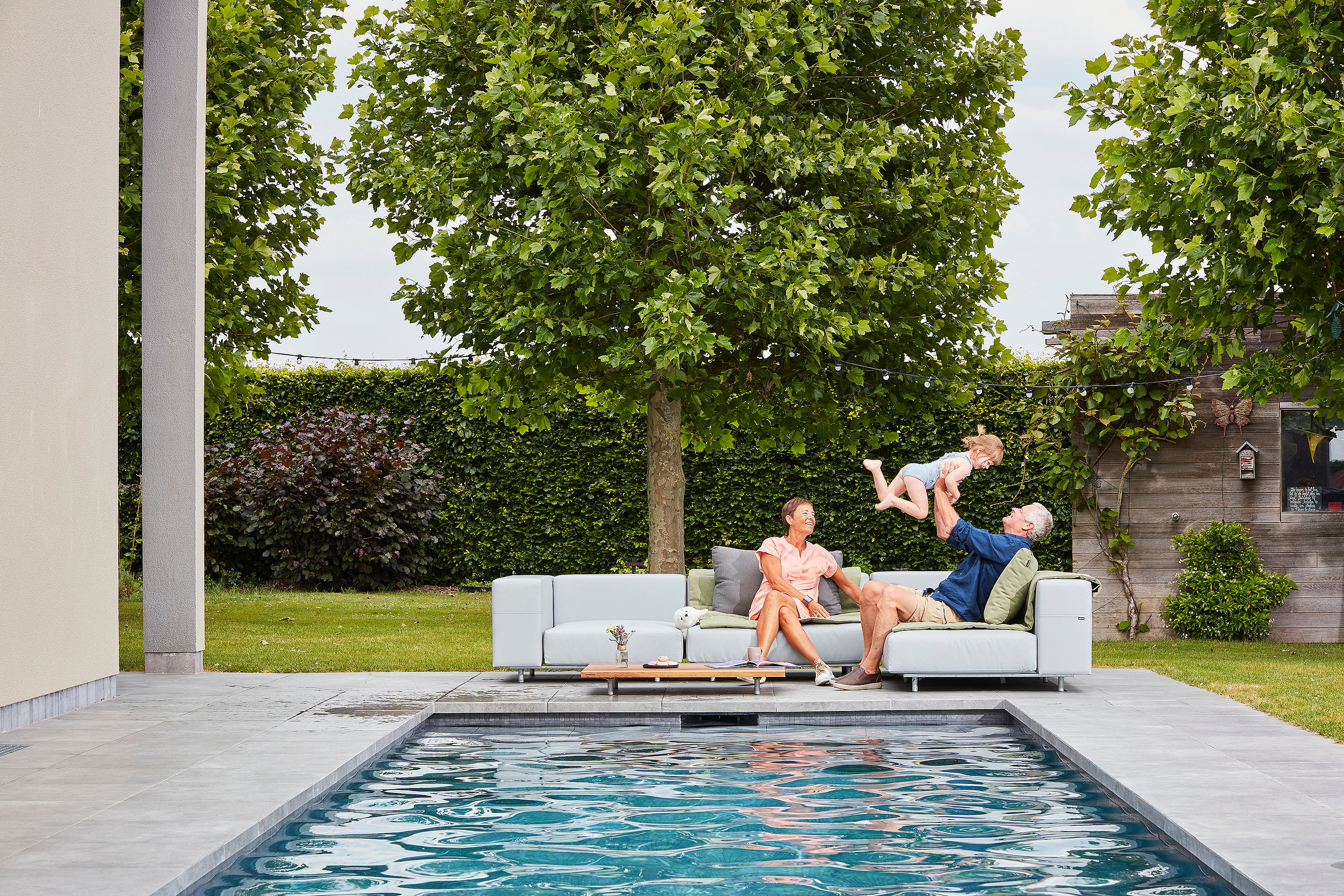 Outdoor-Sofa aus grauem Walross neben einem schmalen Swimmingpool, wo ein älterer Mann ein kleines Kind in die Luft hebt, während eine Frau lächelnd auf der Terrasse sitzt, mit einer grünen Hecke und Bäumen dahinter.
