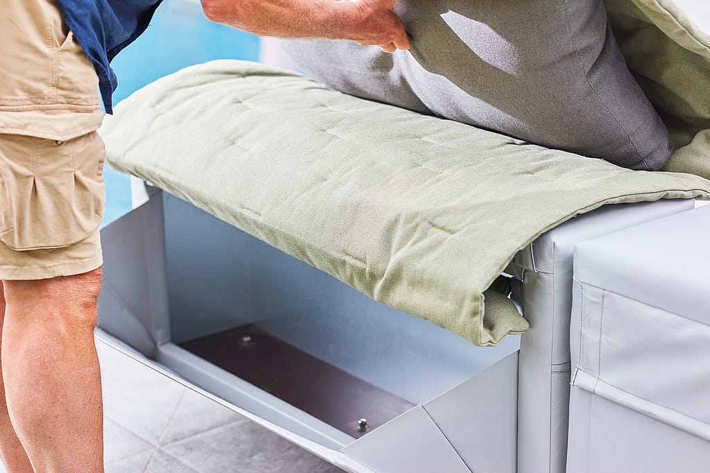 Gros plan d’une personne soulevant le coussin d’un canapé gris Walrus pour révéler le compartiment de rangement intégré en dessous.