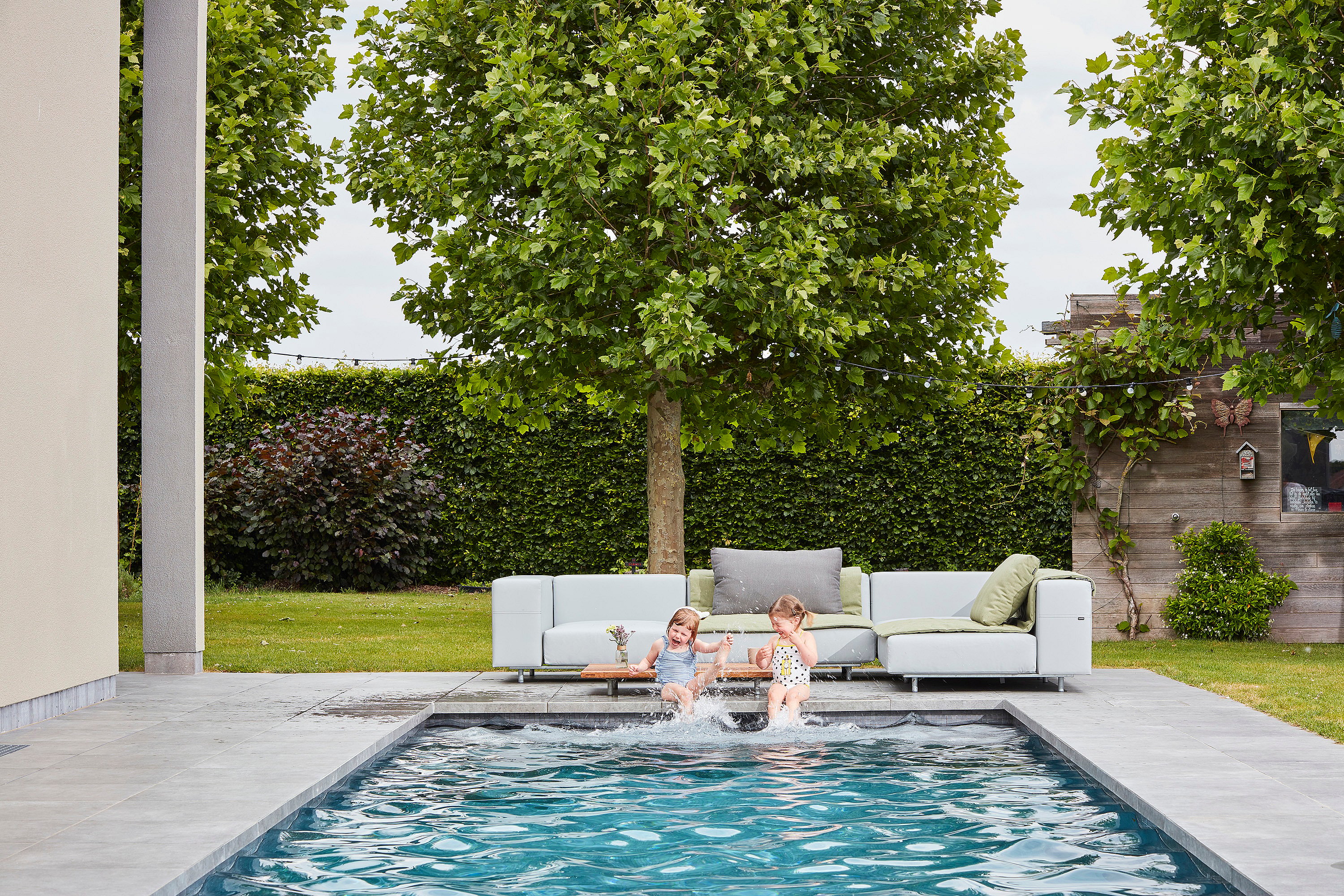 Zwei junge Mädchen in Badeanzügen sitzen am Rande eines schmalen Pools vor einem grauen Walross-Sofa auf einer gepflasterten Gartenterrasse.