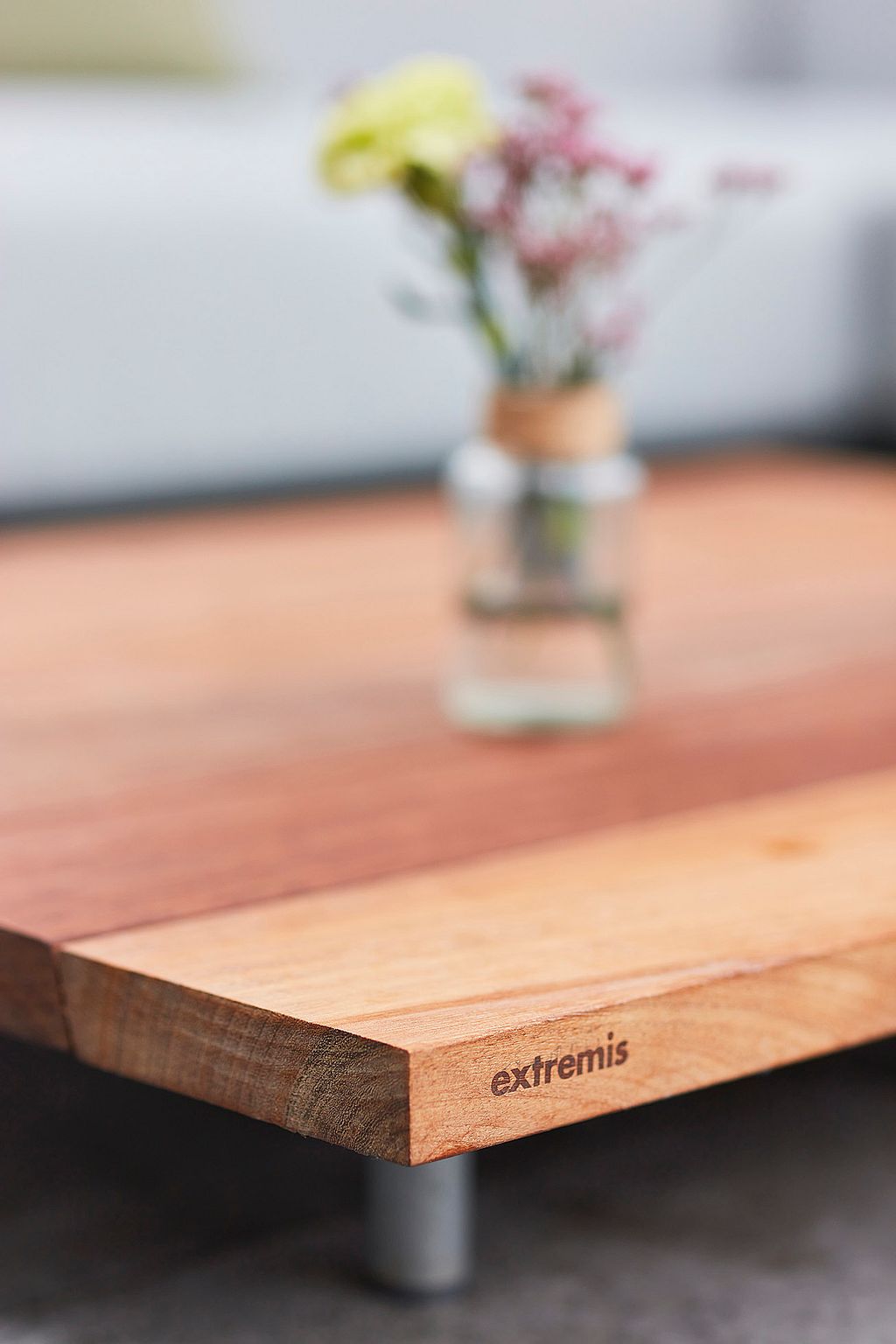 Gros plan d’une table basse en bois pour l’extérieur avec le logo Extremis gravé sur le bord, avec un petit vase de fleurs doucement flouté en arrière-plan.