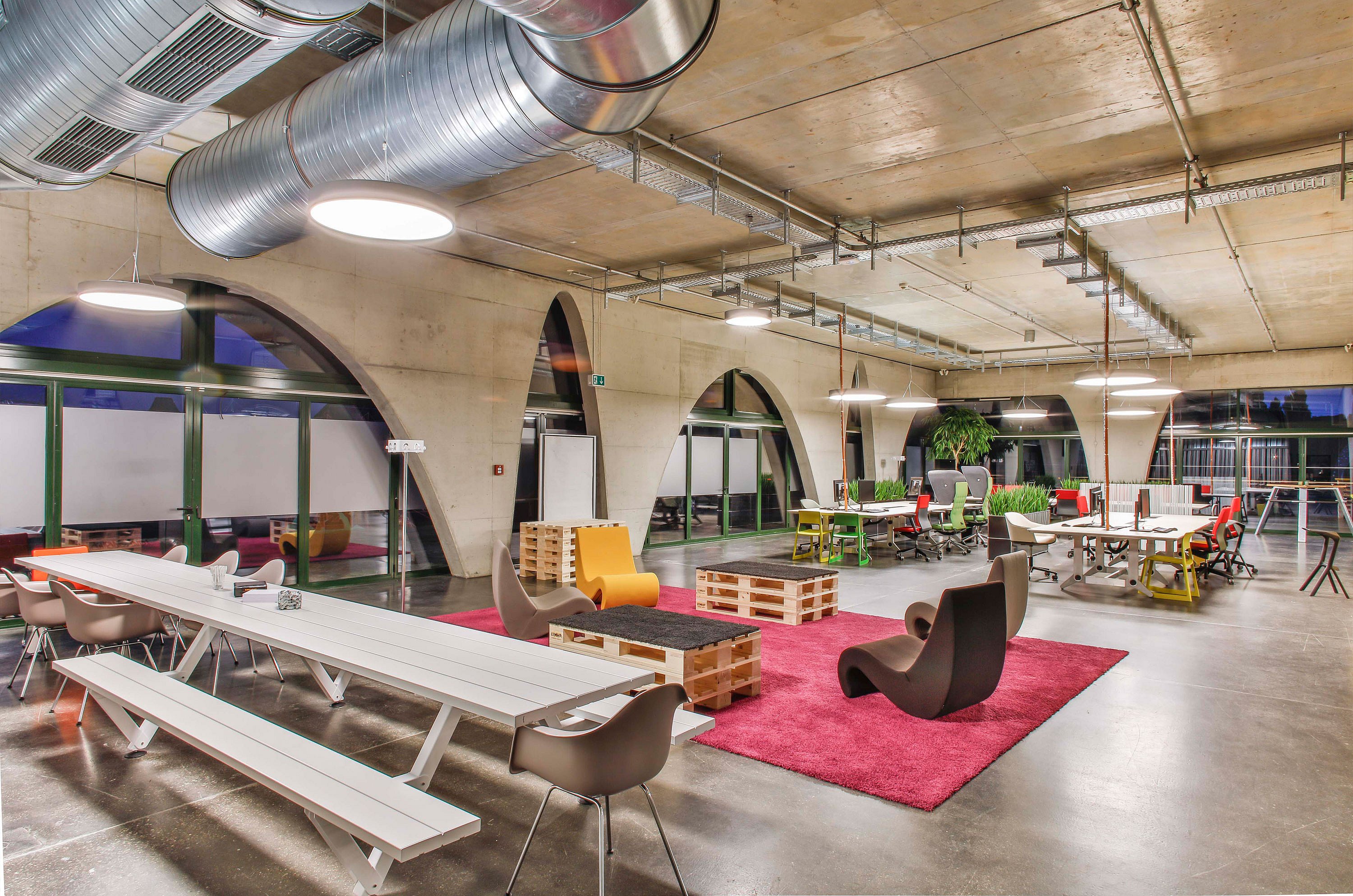 Wide open-plan workspace with a long white Marina picnic table, red rugs, pallet coffee tables, desks, and lounge chairs beneath concrete arches and large circular lights.