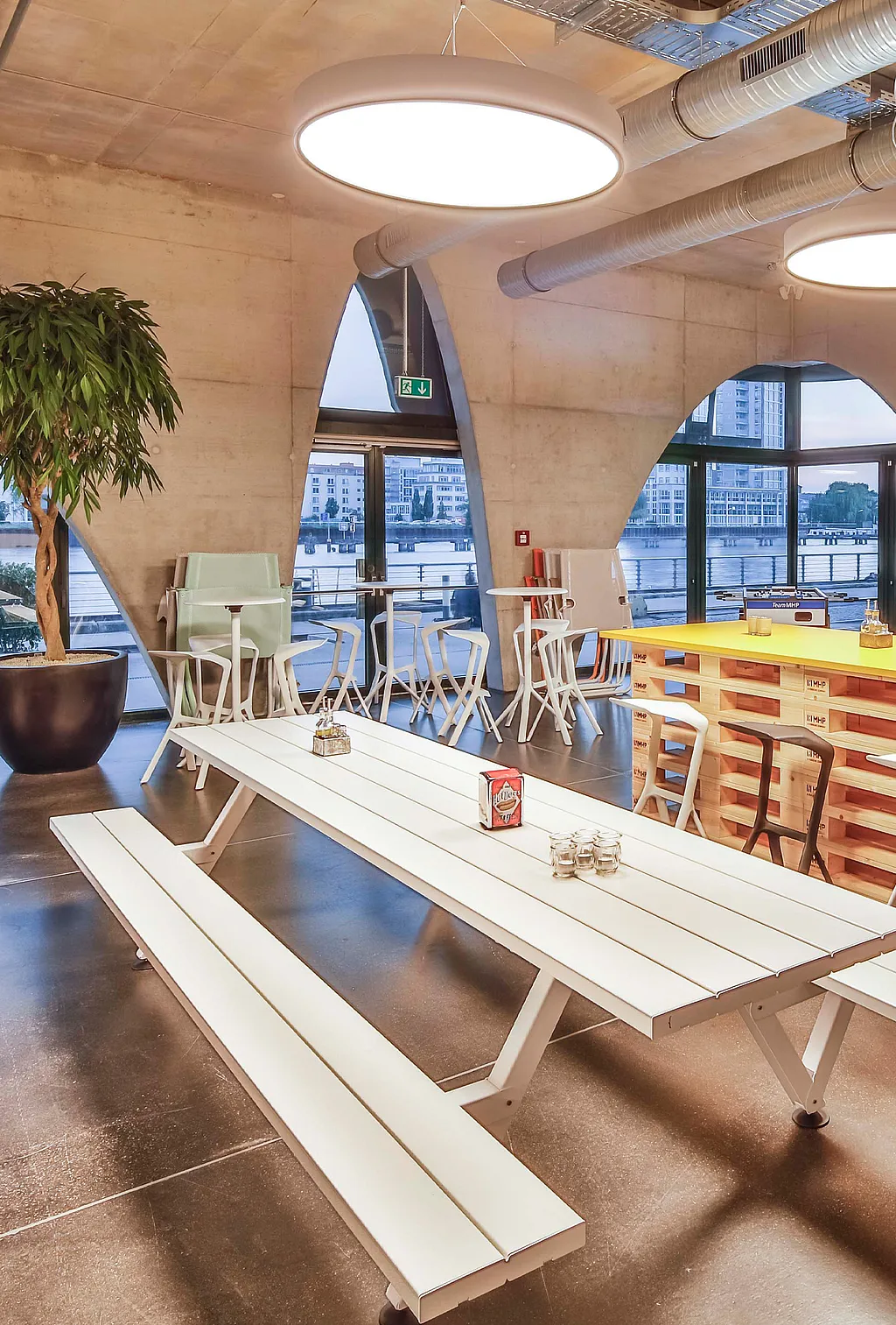 Long white Marina picnic table in an industrial office with exposed concrete arches, round pendant lights, silver ventilation ducts, and riverside windows in the background.