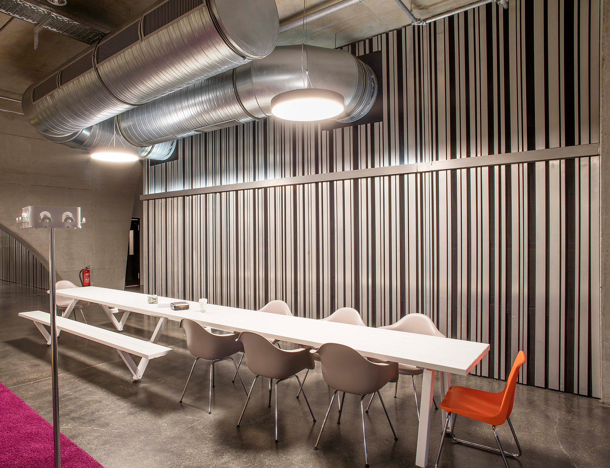 Long white Marina table with bench and chairs beside a tall striped partition wall, under exposed ventilation ducts and round pendant lights in a modern office interior.