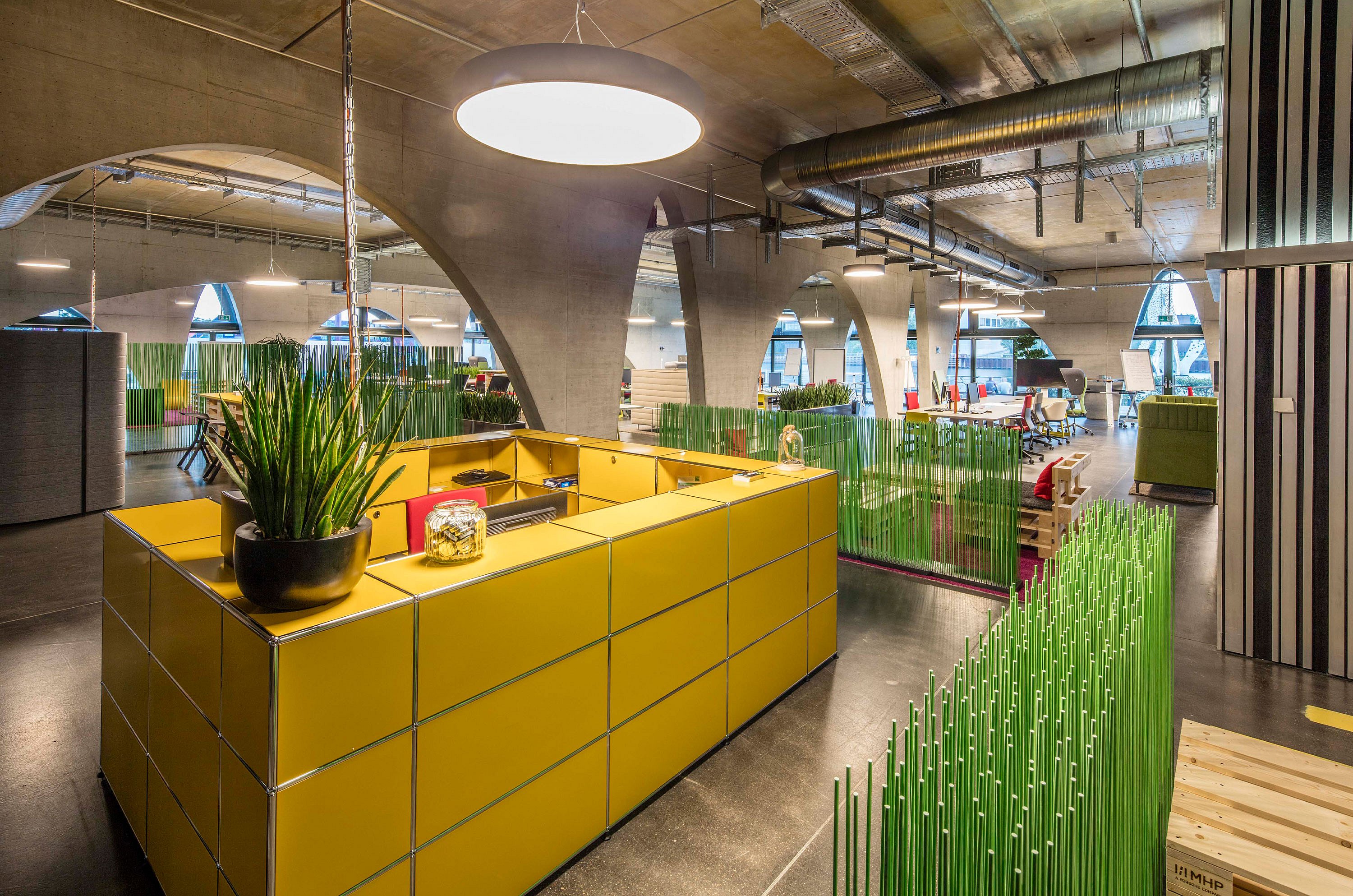 Yellow reception-style counter in a spacious industrial office, with green Sticks partitions dividing work areas beneath exposed ducts, concrete arches, and pendant lights.