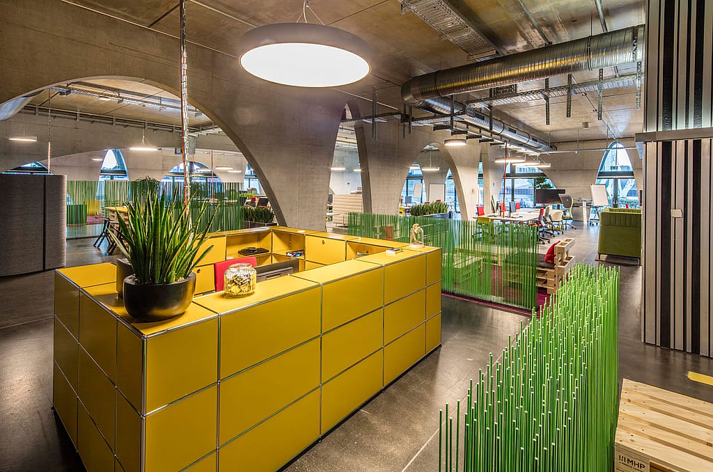 Yellow reception-style counter in a spacious industrial office, with green Sticks partitions dividing work areas beneath exposed ducts, concrete arches, and pendant lights.