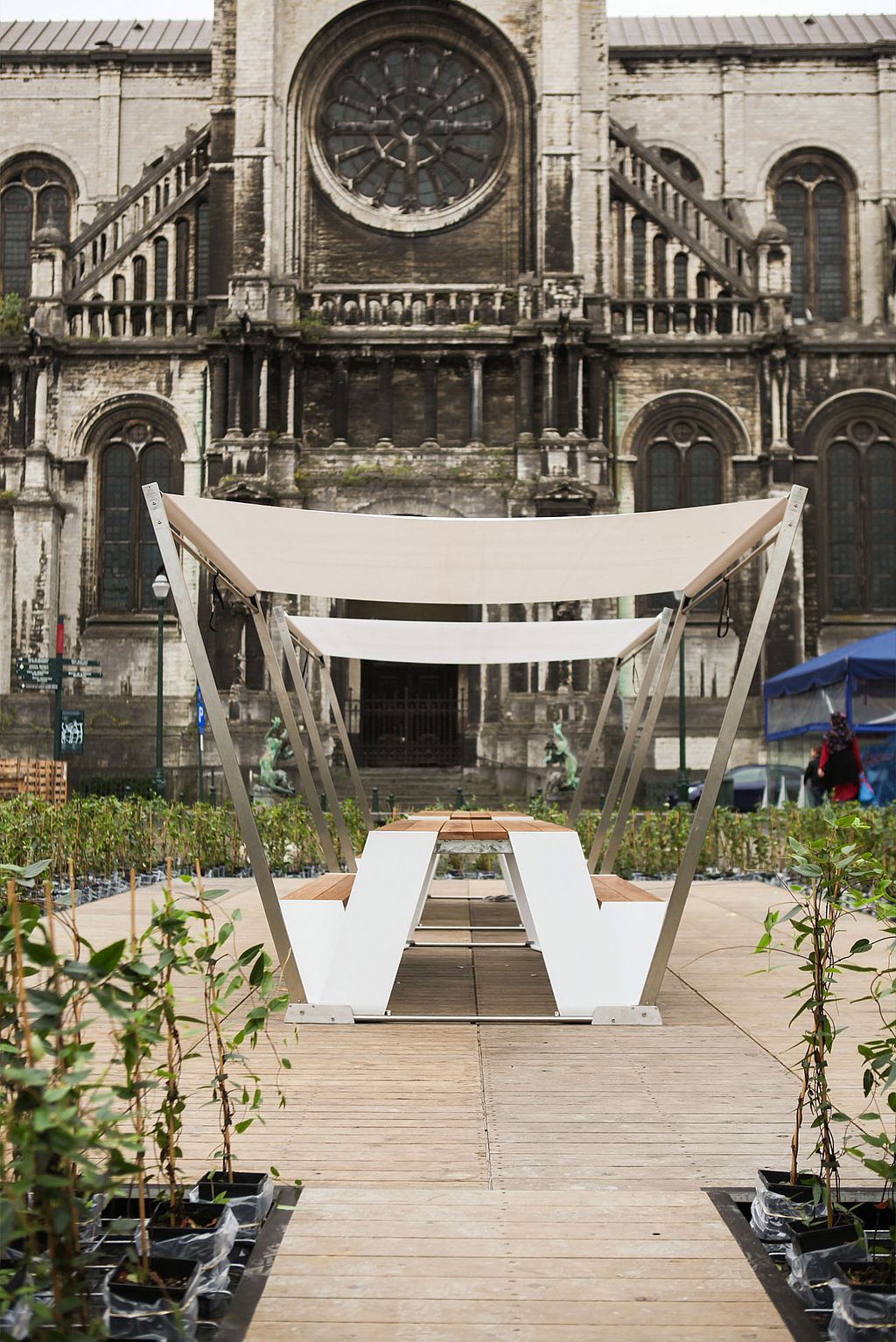 Seitenansicht eines Hopper-Picknicktisches unter geschichtetem weißem Schatten auf einer Holzplattform, ausgerichtet auf die große Fassade und das Rosettenfenster einer historischen Kirche.